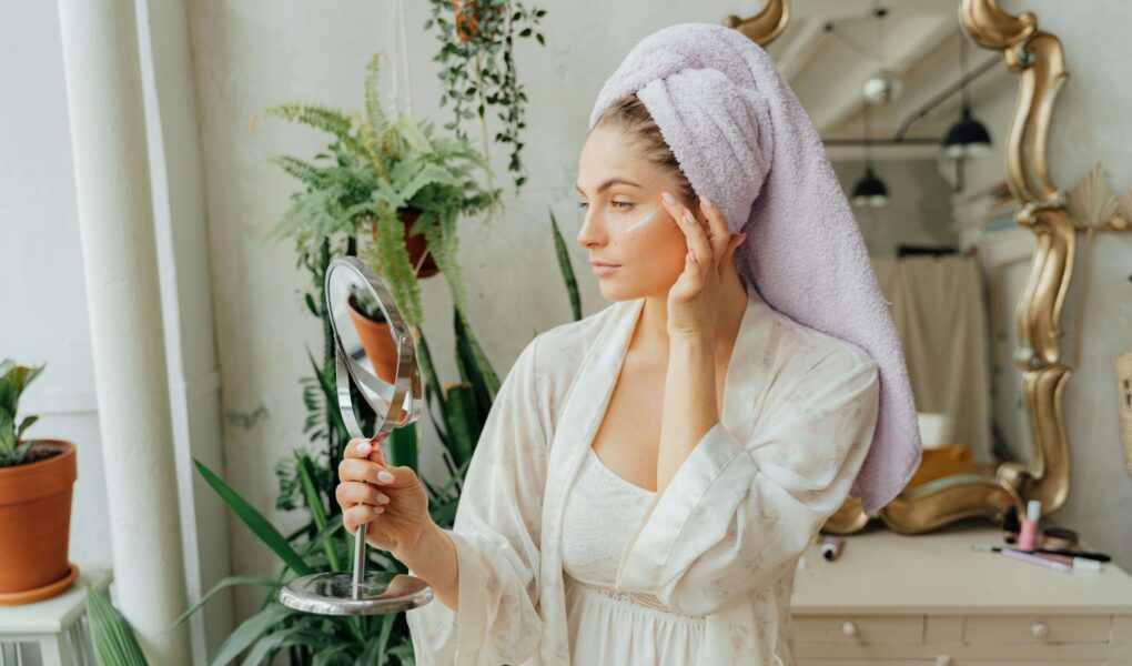 Beautiful woman looking into a hand mirror in the bathroom. best at home facial kit. Self care sunday. diy face mask. diy facial at home. at home facial steps. at home facial steamer. Best facial for glowing skin. how to get radiant skin.