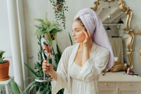 Beautiful woman looking into a hand mirror in the bathroom. best at home facial kit. Self care sunday. diy face mask. diy facial at home. at home facial steps. at home facial steamer. Best facial for glowing skin. how to get radiant skin.