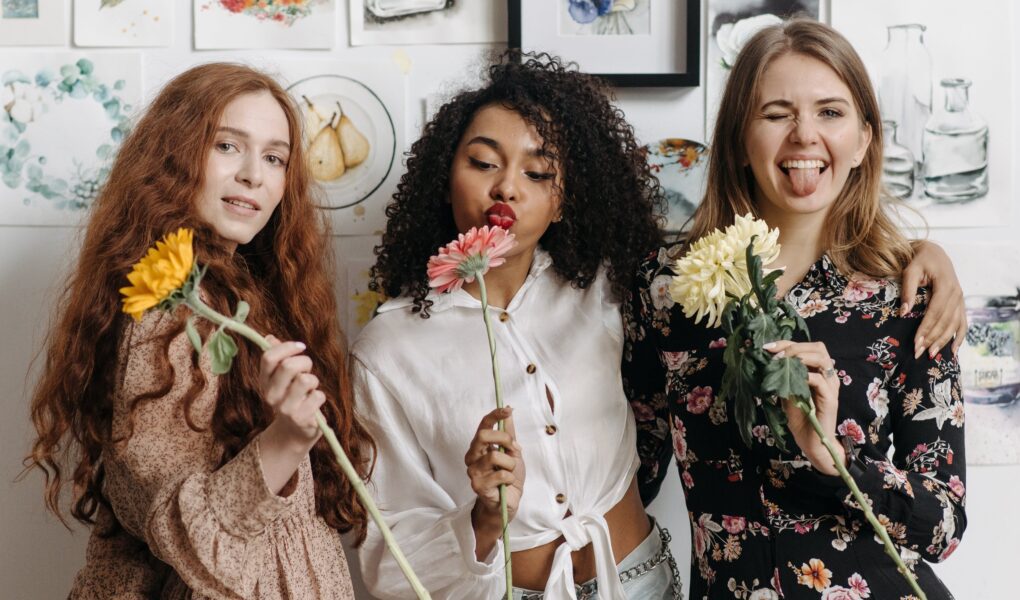 three happy young women holding flowers, smiling and blowing kisses at the camera. galentines dinner ideas. February 13th. galentines day ideas. when is galentines day. girls night cocktails. girls night dinner ideas. girls night snack ideas. girls night in ideas.what to do for girls night. what to do on a girls night. Girl dinner.