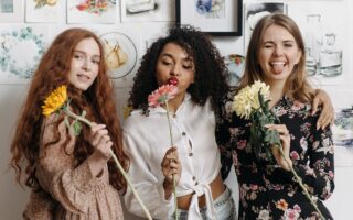 three happy young women holding flowers, smiling and blowing kisses at the camera. galentines dinner ideas. February 13th. galentines day ideas. when is galentines day. girls night cocktails. girls night dinner ideas. girls night snack ideas. girls night in ideas.what to do for girls night. what to do on a girls night. Girl dinner.