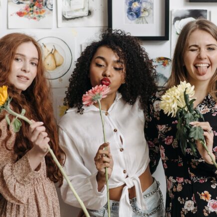 three happy young women holding flowers, smiling and blowing kisses at the camera. galentines dinner ideas. February 13th. galentines day ideas. when is galentines day. girls night cocktails. girls night dinner ideas. girls night snack ideas. girls night in ideas.what to do for girls night. what to do on a girls night. Girl dinner.