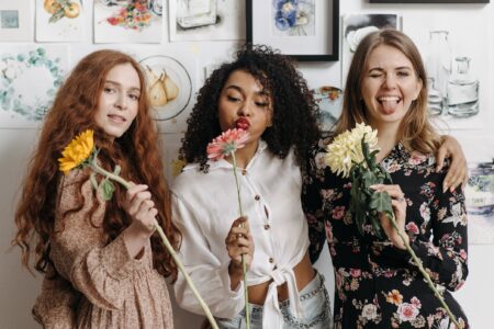 three happy young women holding flowers, smiling and blowing kisses at the camera. galentines dinner ideas. February 13th. galentines day ideas. when is galentines day. girls night cocktails. girls night dinner ideas. girls night snack ideas. girls night in ideas.what to do for girls night. what to do on a girls night. Girl dinner.