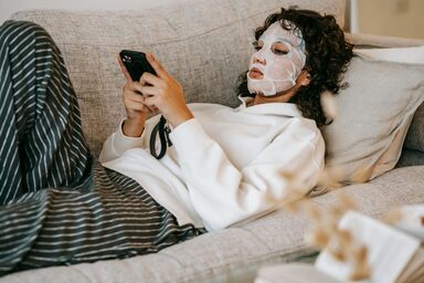 young woman on the couch wearing a sheet mask, on her phone. Hangover breakfast ideas. hangover skin. how to fix a hangover quick. how to treat dehydrated skin. how to fix dehydrated skin. tired when hungover. re energize.