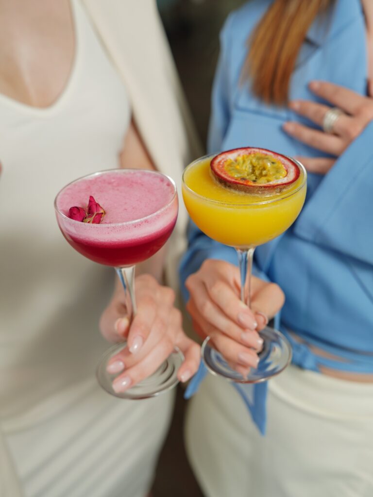 close up of 2 womens hands holding colorful cocktails. galentines dinner ideas. February 13th. galentines day ideas. when is galentines day. girls night cocktails. girls night dinner ideas. girls night snack ideas. girls night in ideas.what to do for girls night. what to do on a girls night. Girl dinner.