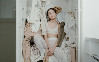 woman sitting in a wardobe with clothes falling out. How to declutter your home. declutter benefits.declutter clothes. how to declutter closet. declutter wardrobe. minimalist wardrobe. how to declutter clothes. how to declutter your closet. emma chamberlain. Underconsumption.