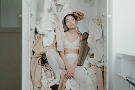 woman sitting in a wardobe with clothes falling out. How to declutter your home. declutter benefits.declutter clothes. how to declutter closet. declutter wardrobe. minimalist wardrobe. how to declutter clothes. how to declutter your closet. emma chamberlain. Underconsumption.