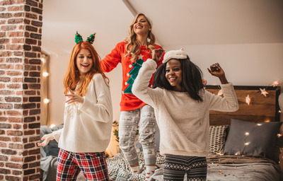 Three young women having Christmas eve pajama party. They are having great time in decorated room, drinking hot chocolate, dancing on the bed. Christmas girls night. Christmas party. Christmas ladies night. holiday girls night. Christmas cocktails. girls night in. holiday party theme. ugly sweater party. 